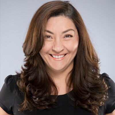 A smiling woman with long, wavy brown hair, embodying the essence of social emotional learning activities, is wearing a black top and standing against a light gray background.