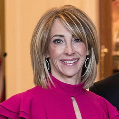 A woman with shoulder-length blonde hair smiles at the camera, confidently displaying her bright pink top and silver earrings. She appears to be in an indoor setting, possibly preparing for an SEL curriculum session with blurred background elements suggesting a dynamic learning environment.