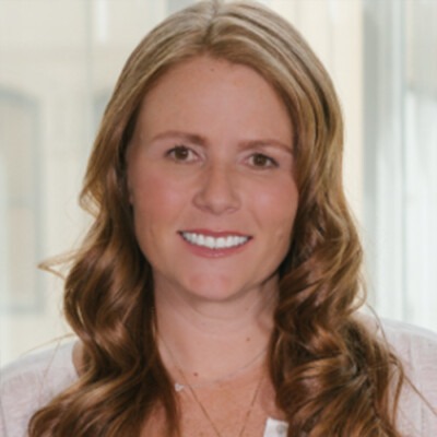 A woman with long, wavy light brown hair smiles at the camera. Wearing a light-colored top, she stands against a soft-focus backdrop featuring windows, embodying the warmth and connection often explored in social emotional learning curriculum.