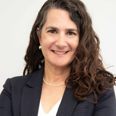 A person with long, curly hair is smiling warmly. They are wearing a dark blazer, a white top, and a pearl necklace, embodying the confidence nurtured through social emotional learning activities. The background is plain and light-colored.