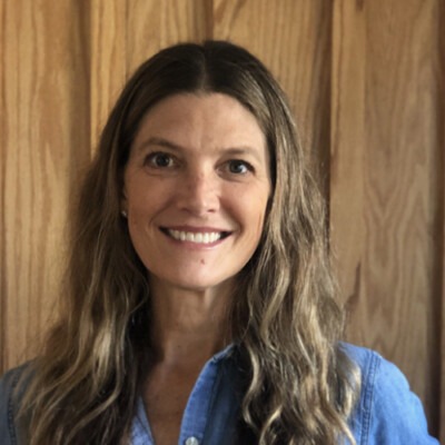 A woman with long, wavy hair smiles at the camera. She is wearing a denim shirt and standing in front of a wooden background, embodying the warmth and creativity often fostered by social emotional learning activities.