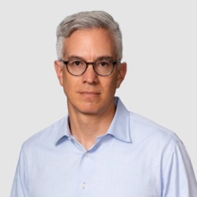 A man with short gray hair and glasses, wearing a light blue button-up shirt, stands with a neutral expression against a plain gray background, embodying the calm and focus often fostered by SEL activities.