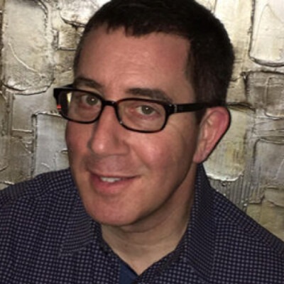 A person with short dark hair and glasses, wearing a dark checkered shirt, smiles at the camera. The background is a textured wall with abstract patterns in shades of gray and beige, adding an artistic touch reminiscent of diversity programs that celebrate individuality.