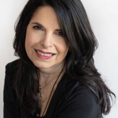A woman with long, dark hair, wearing a black top, smiles at the camera against a plain background, embodying the warmth found in SEL activities.
