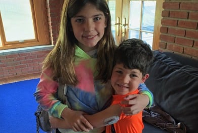 A young girl with long hair in a tie-dye hoodie and a boy in an orange shirt embrace and smile at the camera in a sunlit room, reflecting the warmth of social emotional learning activities. Both wear medical patches on their arms, adding depth to their shared journey.