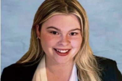 A person with long, light hair smiles at the camera, exuding warmth and openness. They wear a black blazer over a white top against a blue background, embodying the essence of diversity programs in fashion.