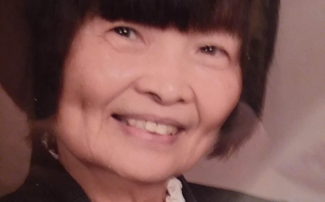 A person with short, dark hair smiles warmly, wearing a dark blazer over a white ruffled blouse. The soft brown background adds an inviting touch, reflecting the inclusive spirit often fostered in diversity programs.
