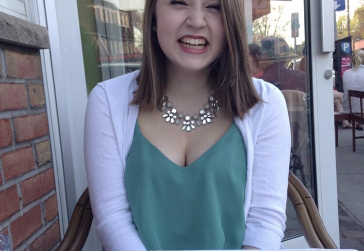 A person smiling while seated at an outdoor table, wearing a white cardigan over a teal top and a statement necklace, embodies the joy often inspired by engaging in social emotional learning activities. A knife and fork rest nearby, with a brick wall and street visible in the background.