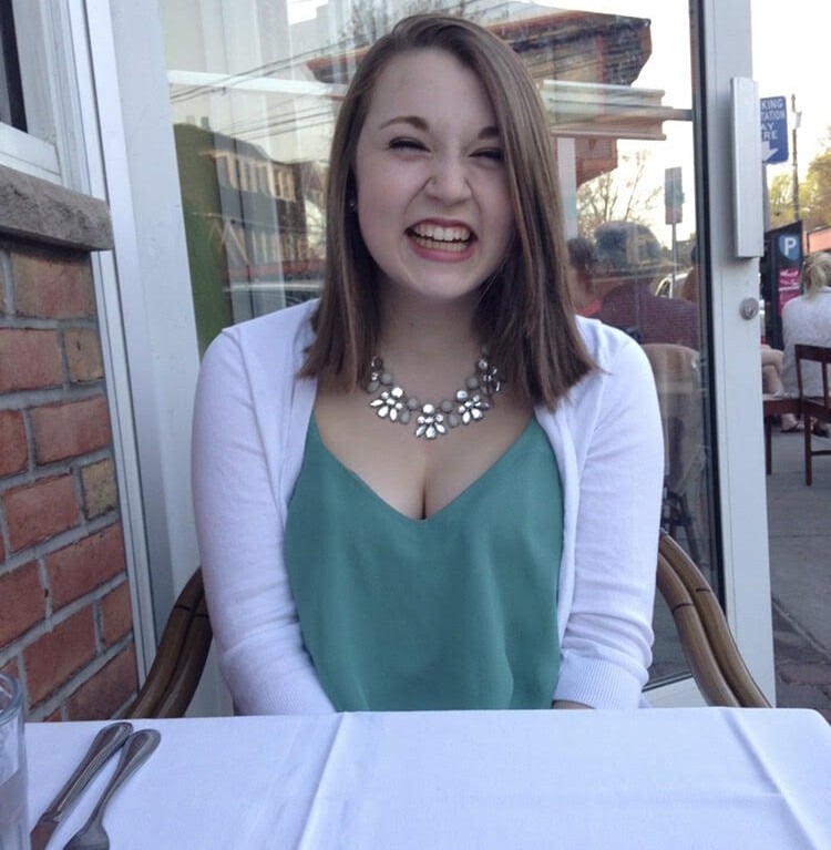 A person smiling while seated at an outdoor table, wearing a white cardigan over a teal top and a statement necklace, embodies the joy often inspired by engaging in social emotional learning activities. A knife and fork rest nearby, with a brick wall and street visible in the background.