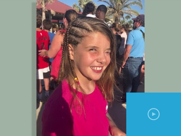 A young girl with braided hair smiles while standing outdoors, embracing the spirit of diversity programs. She is wearing a bright pink T-shirt, and people and palm trees bask in the sunny background.