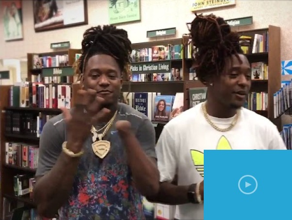 Two men with dreadlocks and necklaces stand in a bookstore, embodying the spirit of diversity programs. They are smiling, surrounded by shelves filled with books. One wears a patterned shirt, the other a white shirt. A blue play button in the corner suggests a video thumbnail.