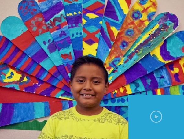 A smiling boy stands in front of a colorful peacock feather mural made of painted paper. The feathers, embodying diversity, are decorated with vibrant patterns in blue, red, yellow, and purple tones. A play button icon is overlaid in the bottom right corner, introducing social emotional learning activities.