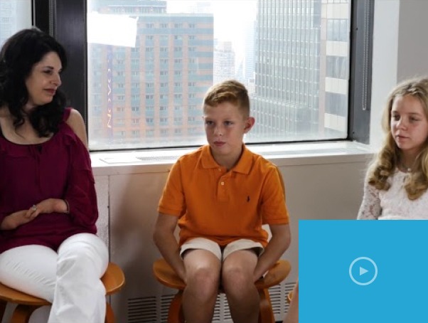 A woman and two children sit on stools in a room with large windows showcasing city skyscrapers, engaged in SEL activities. The woman wears a maroon top, the boy an orange shirt, and the girl a white top. A play button icon is visible in the lower right corner.