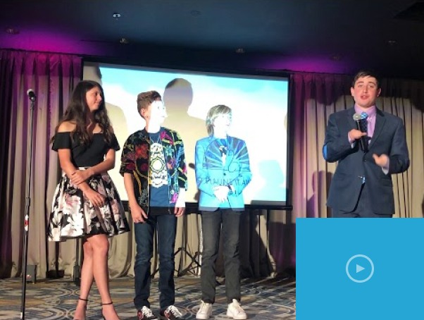 A woman and three young men stand on a stage under spotlights. A microphone is in front of them, and a large screen is partially visible in the background. They appear to be presenting diversity programs at a formal event or presentation.