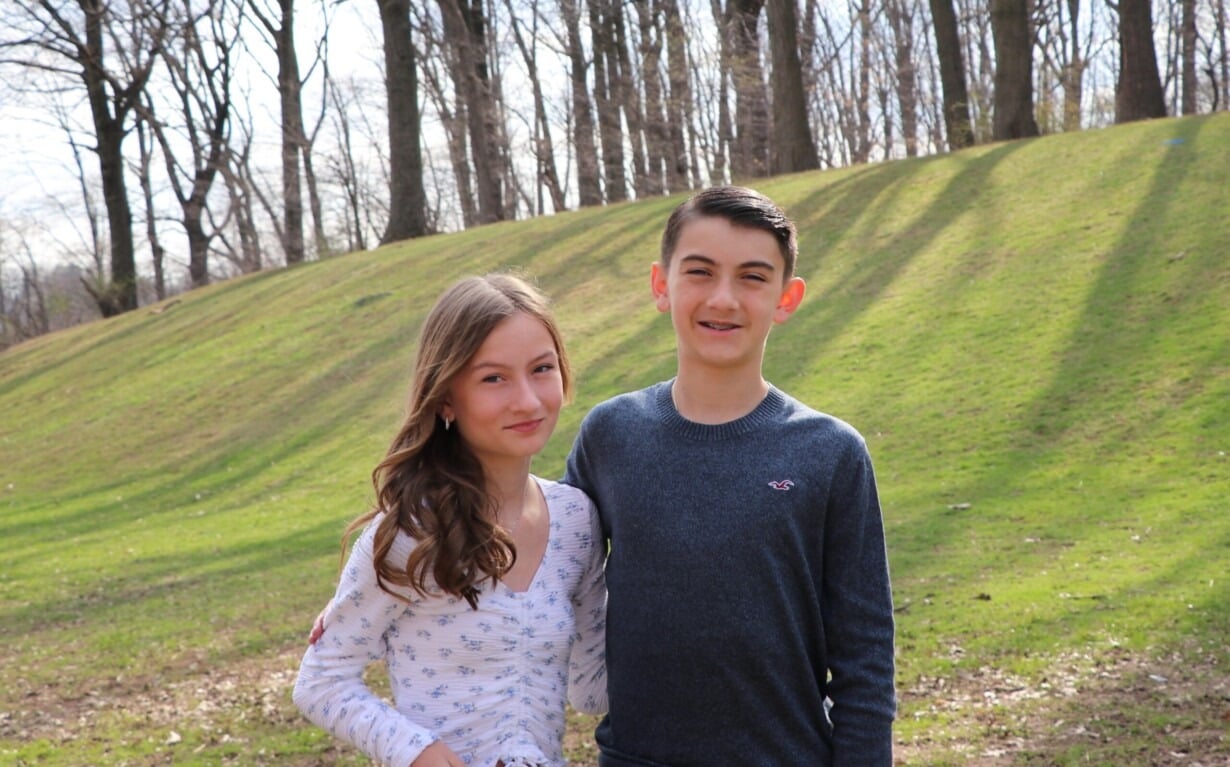Two teenagers stand together outdoors on a sunny day, the girl in a white patterned shirt and the boy in a gray sweater, embodying the spirit of SEL curriculum. They smile against a backdrop of grassy hills and bare trees, embracing life's simple joys.