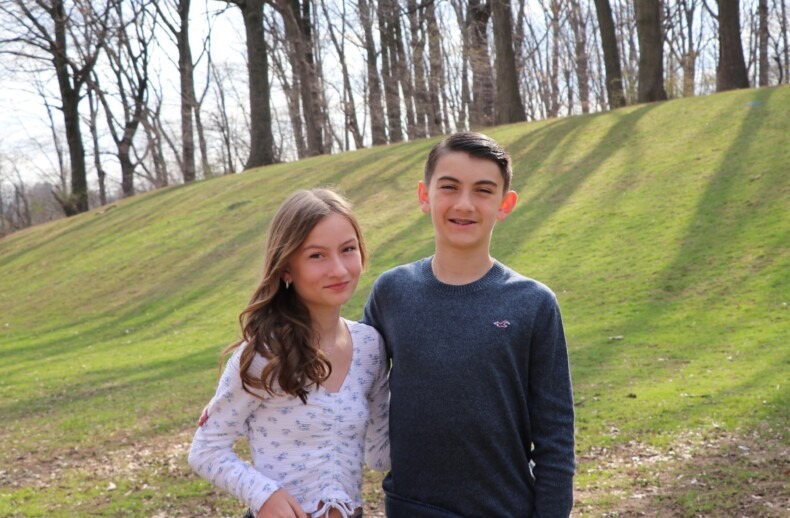 Two teenagers stand together outdoors on a sunny day, the girl in a white patterned shirt and the boy in a gray sweater, embodying the spirit of SEL curriculum. They smile against a backdrop of grassy hills and bare trees, embracing life's simple joys.