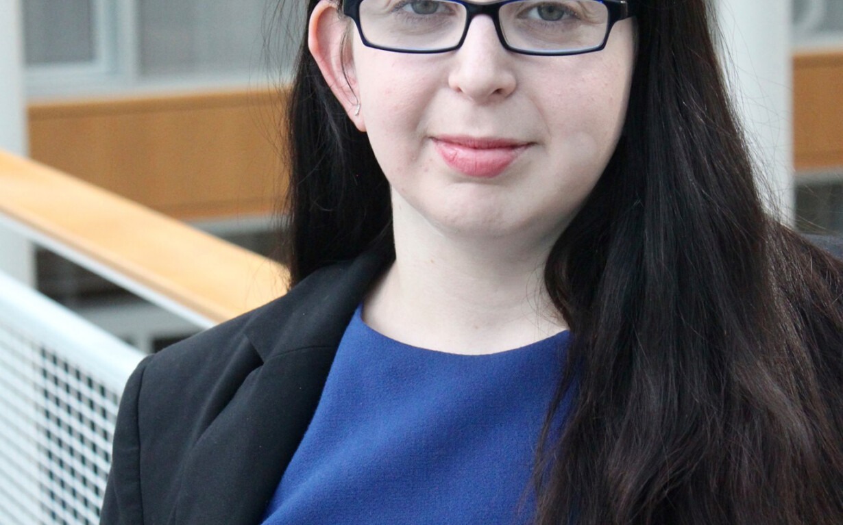 A person with long dark hair, wearing glasses, a blue dress, and a black blazer, is standing indoors. The modern interior, with wooden panels and glass windows, creates an ideal backdrop for discussions on social-emotional learning activities.