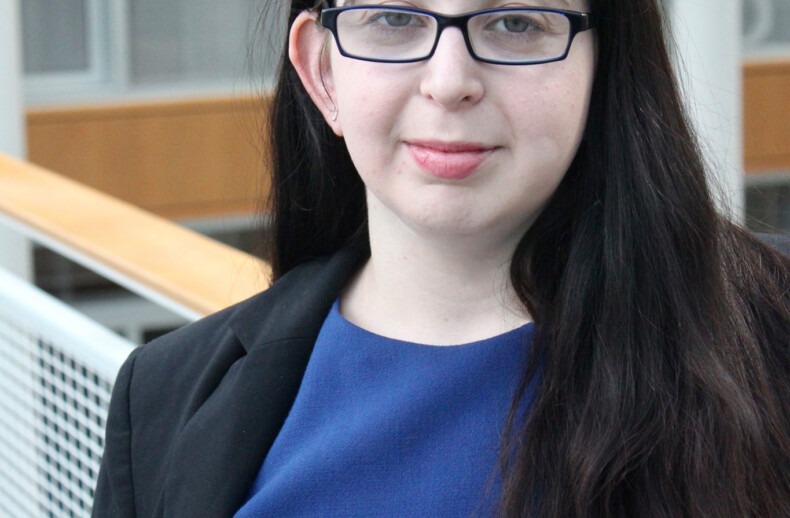 A person with long dark hair, wearing glasses, a blue dress, and a black blazer, is standing indoors. The modern interior, with wooden panels and glass windows, creates an ideal backdrop for discussions on social-emotional learning activities.