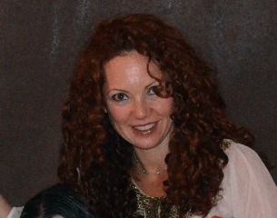A person with curly red hair is smiling at the camera, wearing a light-colored top with decorative details. The background is dark and blurred, reminiscent of get-to-know-you activities for students that foster connection and warmth in SEL settings.