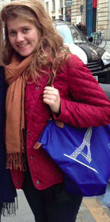 A person with long hair wearing a red jacket and brown scarf stands on a city street, embracing the diversity programs of urban life while holding a blue bag adorned with a white Eiffel Tower design.