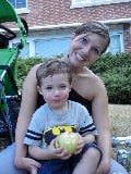 A woman smiling while holding a young boy on her lap, engaging in social emotional learning activities. The boy clutches a yellow apple as they sit on the grass in front of a brick building, with a stroller nearby.