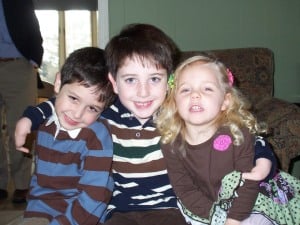 EthanCharlieSavanna Three children are sitting closely together, smiling warmly as if they just completed fun get-to-know-you activities for students. Two boys on the left wear striped shirts, while a girl on the right flaunts her blonde hair and pink clip. Indoors, a window and chair complete the cozy scene.