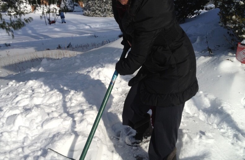 A person dressed in black winter clothing and a fur hat is smiling while shoveling snow, embracing a moment of personal growth akin to social emotional learning activities. They are outside in a snowy landscape with trees in the background.