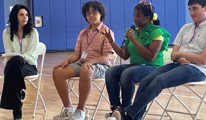 A group of four people seated on chairs in a gymnasium participates in an SEL curriculum session. One person speaks into a microphone as the others listen attentively, all wearing event lanyards and casual attire, fostering an inclusive and engaging atmosphere.