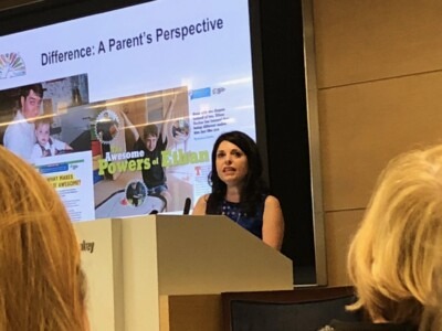 A woman speaks at a podium with a presentation slide behind her titled "Difference: A Parent's Perspective," featuring images and text about a child named Ethan. The talk highlights SEL curriculum and its impact, while audience members listen intently in the foreground.