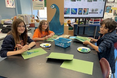 Three students sit at a table in an art classroom, painting with green and blue shades. Engaged in social emotional learning activities, they focus intently amid a backdrop of art supplies and a peacock poster, fostering creativity and connection through their shared artistic journey.