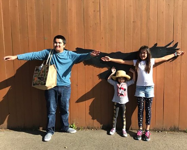 A man and two children stand against a wooden wall with a painted bird silhouette, their arms outstretched to mimic its wings. The man holds a patterned bag, while the child in the middle wears a large straw hat, reflecting social emotional learning activities through creative expression.