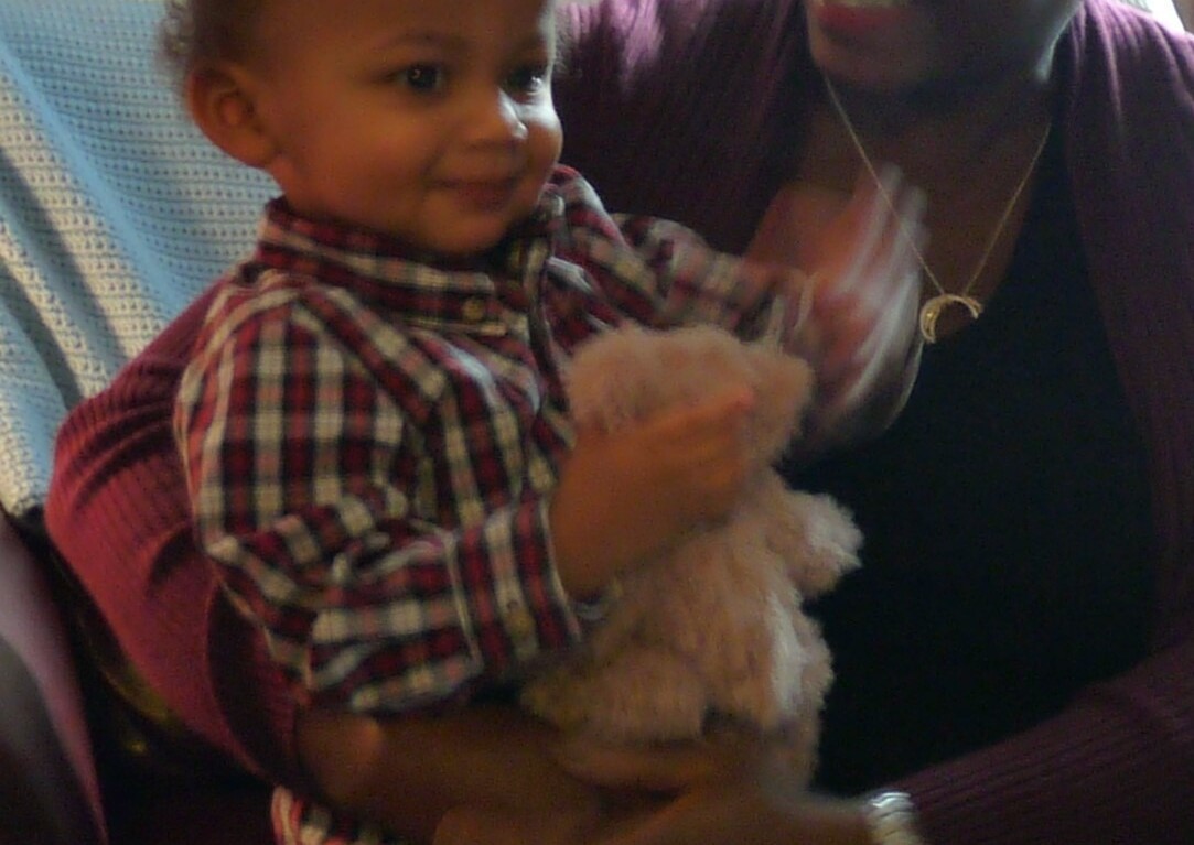 A smiling adult holds a young child on their lap during a warm moment of social emotional learning activities. The child, wearing a plaid shirt and clutching a stuffed toy, sits indoors surrounded by floral curtains and a blue blanket in the background.