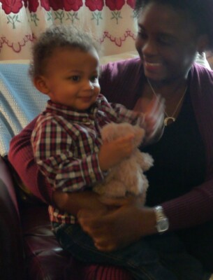 A smiling adult holds a young child on their lap during a warm moment of social emotional learning activities. The child, wearing a plaid shirt and clutching a stuffed toy, sits indoors surrounded by floral curtains and a blue blanket in the background.