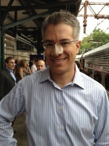 A man with short gray hair, wearing glasses and a striped shirt, smiles at a train station, perhaps on his way to an engaging SEL activity. He has a bandage on his nose. Other people and a train are visible in the background.
