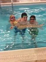 Three children are in a swimming pool. Two boys are holding a girl who wears goggles and a swimsuit as part of their get-to-know-you activities for students. They are smiling and enjoying their time in the water, fostering connection and camaraderie.
