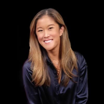 A woman with long, light brown hair is smiling, wearing a dark, silky blouse. She is set against a simple black background, reminiscent of the warmth and connection fostered by social emotional learning activities.