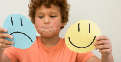 A child with curly hair and a pensive expression holds two paper circles—a blue one with a sad face and a yellow one with a happy face—against a neutral background, embodying the essence of an SEL curriculum activity that helps young minds explore emotions.