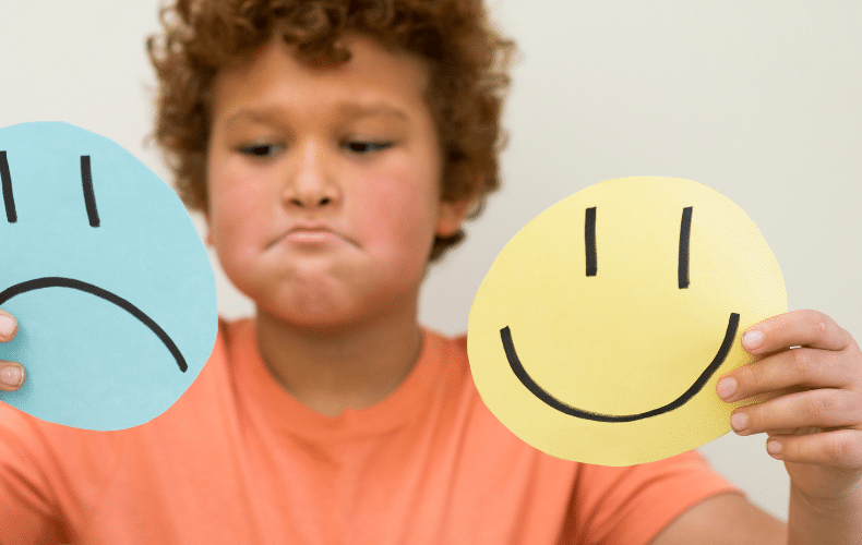A child with curly hair and a pensive expression holds two paper circles—a blue one with a sad face and a yellow one with a happy face—against a neutral background, embodying the essence of an SEL curriculum activity that helps young minds explore emotions.
