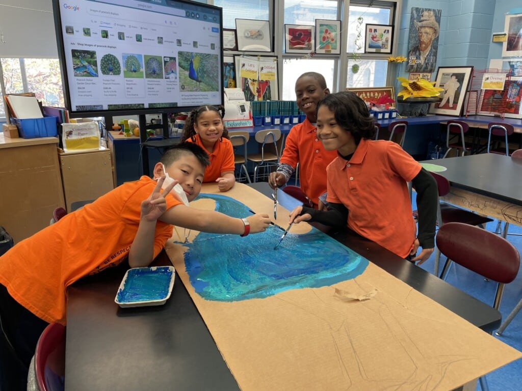 Four children are painting a large sheet of paper with blue paint in a colorful classroom as part of their social-emotional learning activities. They are smiling and wearing matching orange shirts. Art supplies and projects fill the vibrant backdrop, reflecting the joy of creativity and teamwork.