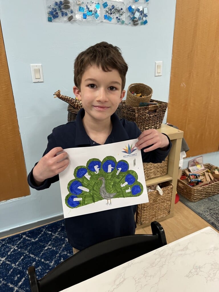 A young boy smiles while holding up a drawing of a peacock with colorful feathers, a perfect project for the classroom's social emotional learning curriculum. In the background, storage baskets and art supplies are visible, enhancing this creative educational setting.