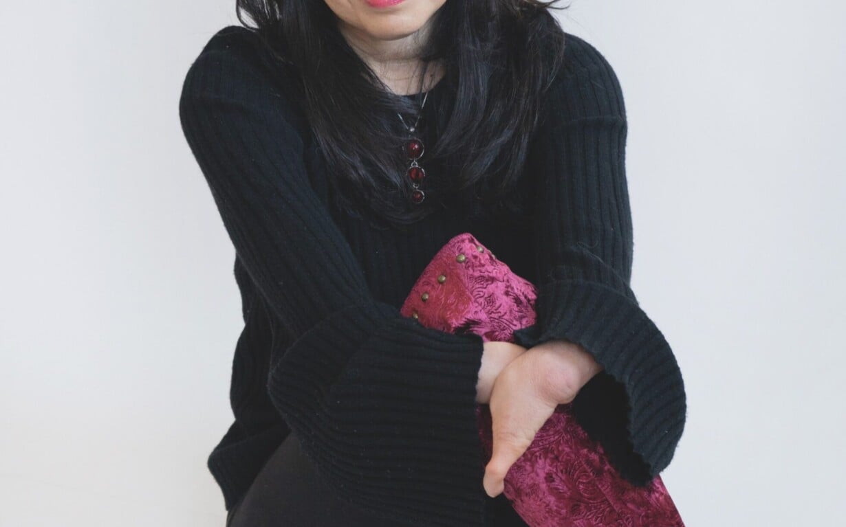 A woman with long, dark hair sits against a white background, smiling softly. Dressed in a black sweater, she holds her crossed legs while sporting maroon, textured boots. Her presence exudes warmth, reminiscent of engaging social emotional learning activities that foster connection.
