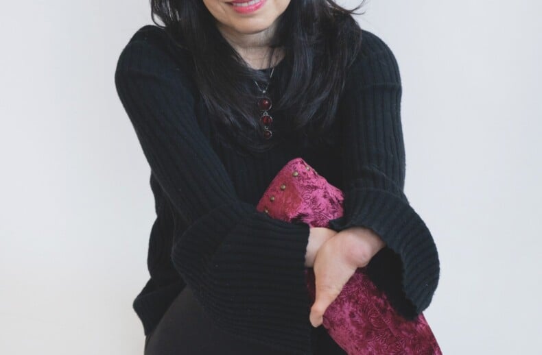 A woman with long, dark hair sits against a white background, smiling softly. Dressed in a black sweater, she holds her crossed legs while sporting maroon, textured boots. Her presence exudes warmth, reminiscent of engaging social emotional learning activities that foster connection.