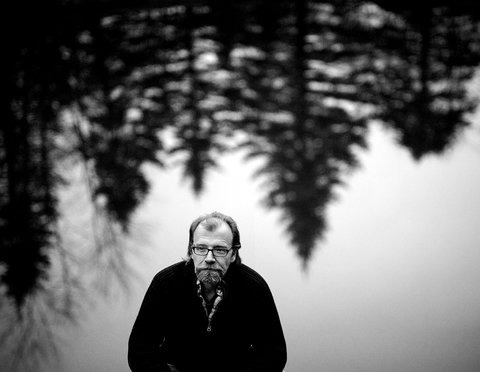 A black and white photo of a man standing by a calm lake with a reflection of trees evokes an air of introspection, much like SEL activities that foster emotional growth. The serene, misty atmosphere with towering evergreens enhances this peaceful scene.