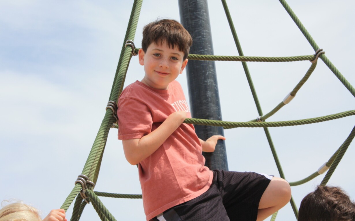 A child wearing a pink T-shirt and black shorts is climbing a rope structure at a playground, showcasing the confidence fostered by social emotional learning curriculum. The sky is clear and sunny.