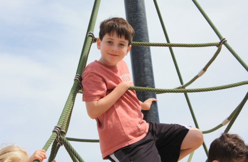 A child wearing a pink T-shirt and black shorts is climbing a rope structure at a playground, showcasing the confidence fostered by social emotional learning curriculum. The sky is clear and sunny.
