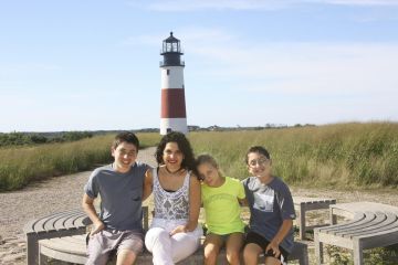 A group of four people sits on a curved bench in front of a lighthouse, enjoying the clear sky and tall grass. Their smiles reflect the joy and connection encouraged by social emotional learning activities, with one adult guiding three children peacefully.