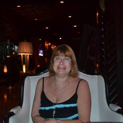A woman with glasses and shoulder-length hair sits in a black and white chair, smiling. She wears a black tank top with blue stripes. The dimly lit background, enhanced with warm light fixtures and a decorative lamp, sets the perfect mood for SEL activities that foster connection and understanding.