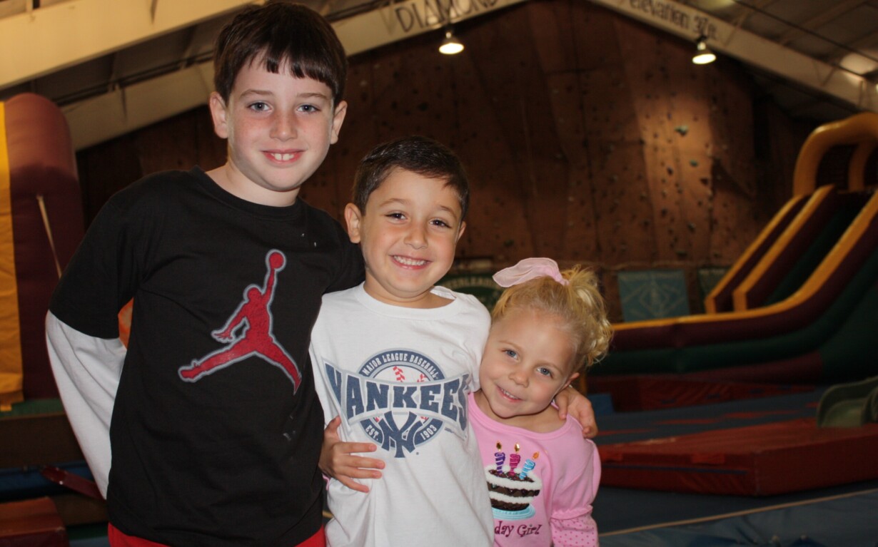 Three children smiling and posing in an indoor play area, enjoying a day filled with fun SEL activities. The boy on the left wears a black T-shirt, the boy in the middle sports a white Yankees shirt, and the girl on the right has a pink shirt with a birthday cake print. Inflatable slides are in the background.