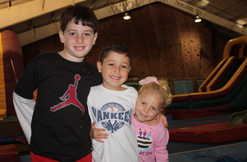Three children smiling and posing in an indoor play area, enjoying a day filled with fun SEL activities. The boy on the left wears a black T-shirt, the boy in the middle sports a white Yankees shirt, and the girl on the right has a pink shirt with a birthday cake print. Inflatable slides are in the background.