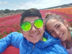 Two children smiling in a selfie, embracing the joy of SEL activities. The boy sports reflective sunglasses and a blue jacket, while the girl has her hair tied up. They enjoy a field full of red and orange flowers under a clear sky, capturing moments of sunshine and friendship.